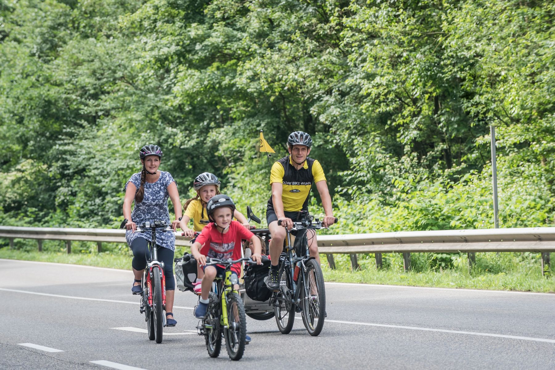 Four people, two adults and two children, ride bicycles on a road bordered by a metal guardrail and lush green trees. All are wearing helmets and casual clothes, and the weather appears clear and pleasant.